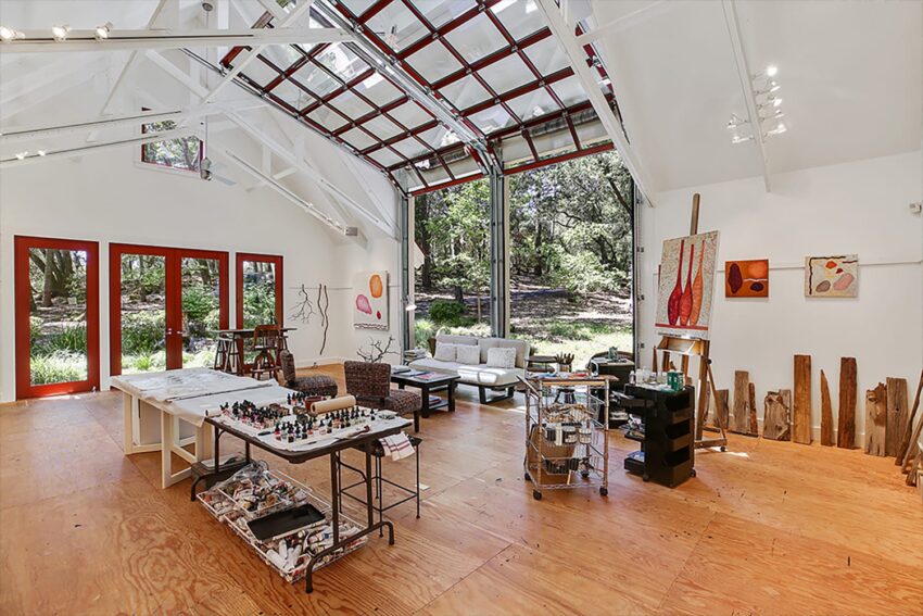 Spacious art studio with high ceilings, colorful paintings, open doors, and a view of a lush forest outside.