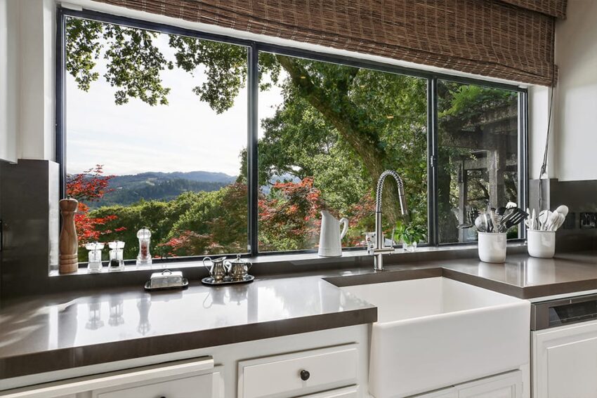 Kitchen with white cabinets, large window view of lush trees, and countertop items like a kettle and pepper grinder.