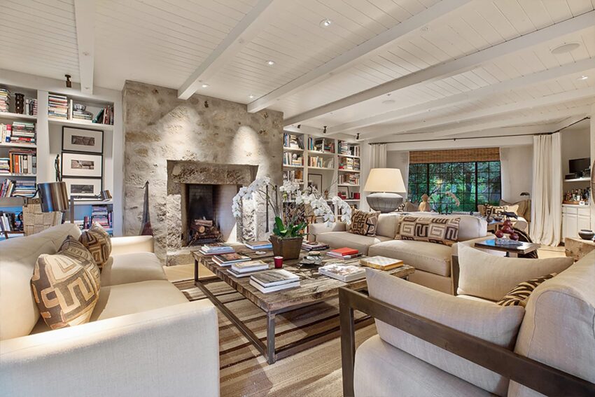 Cozy living room with stone fireplace, wooden ceiling beams, beige sofas, patterned pillows, and bookshelves.