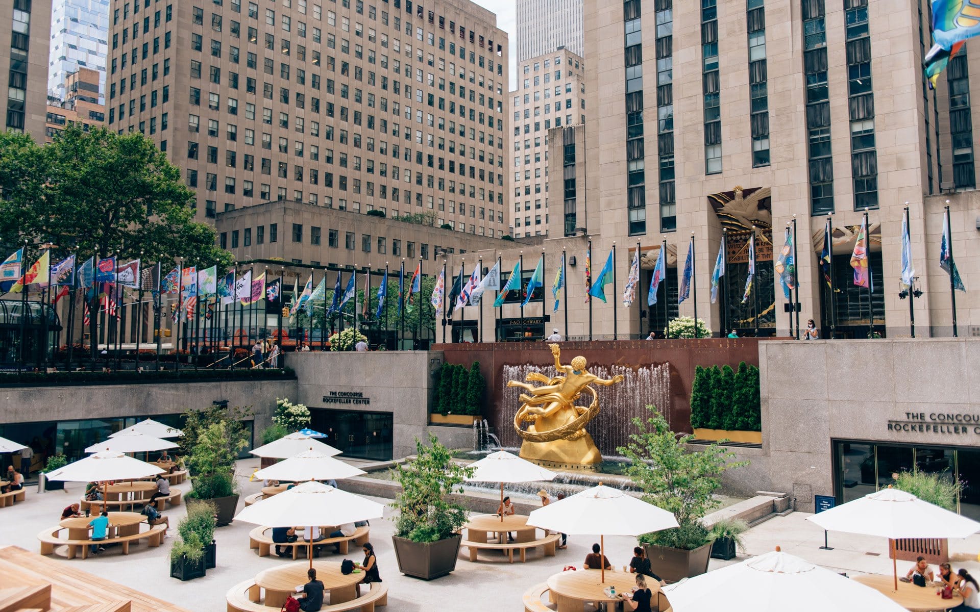 Outdoor seating area with umbrellas and a golden statue surrounded by flags and tall buildings in a bustling city plaza.