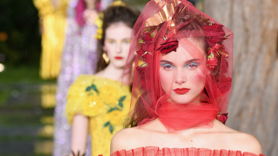 Model in red dress and veil walks runway with models in colorful dresses blurred in background at fashion show.