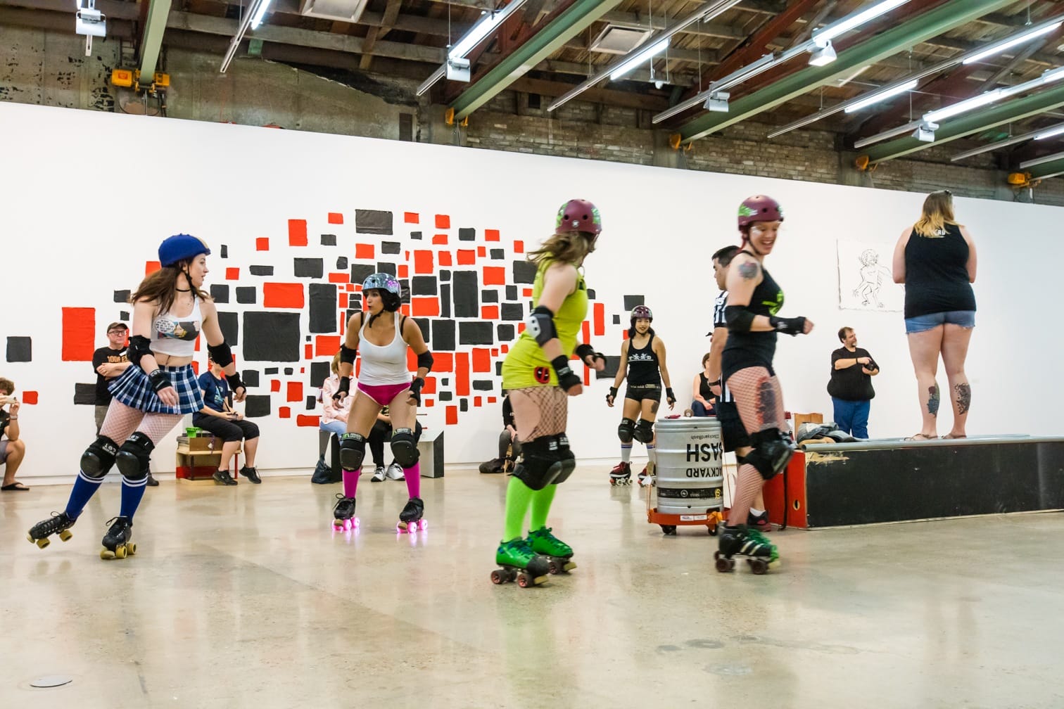 Roller derby skaters in gear practicing indoors with a modern art mural in the background and spectators watching.