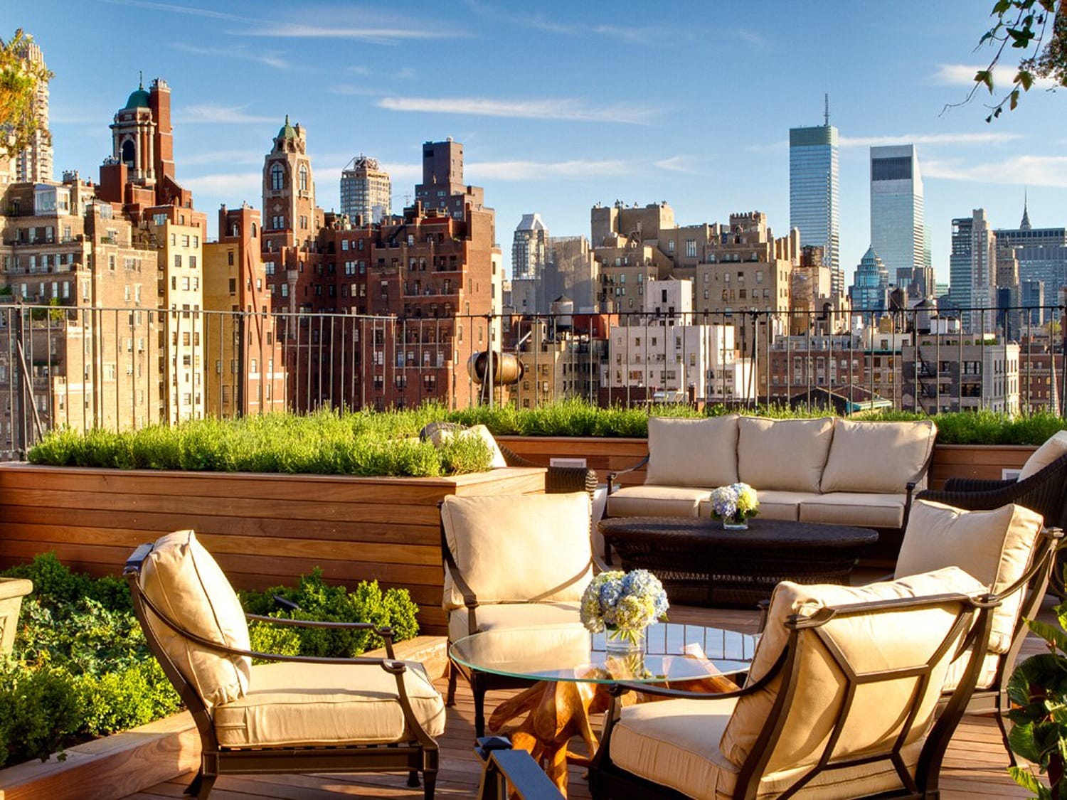 Rooftop patio with seating and plants overlooking a city skyline with tall buildings under a clear blue sky.