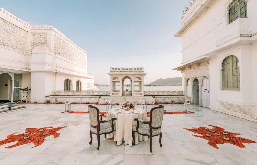 Elegant outdoor dining setup on a palace terrace with ornate architecture and lake view, decorated with flower petals.