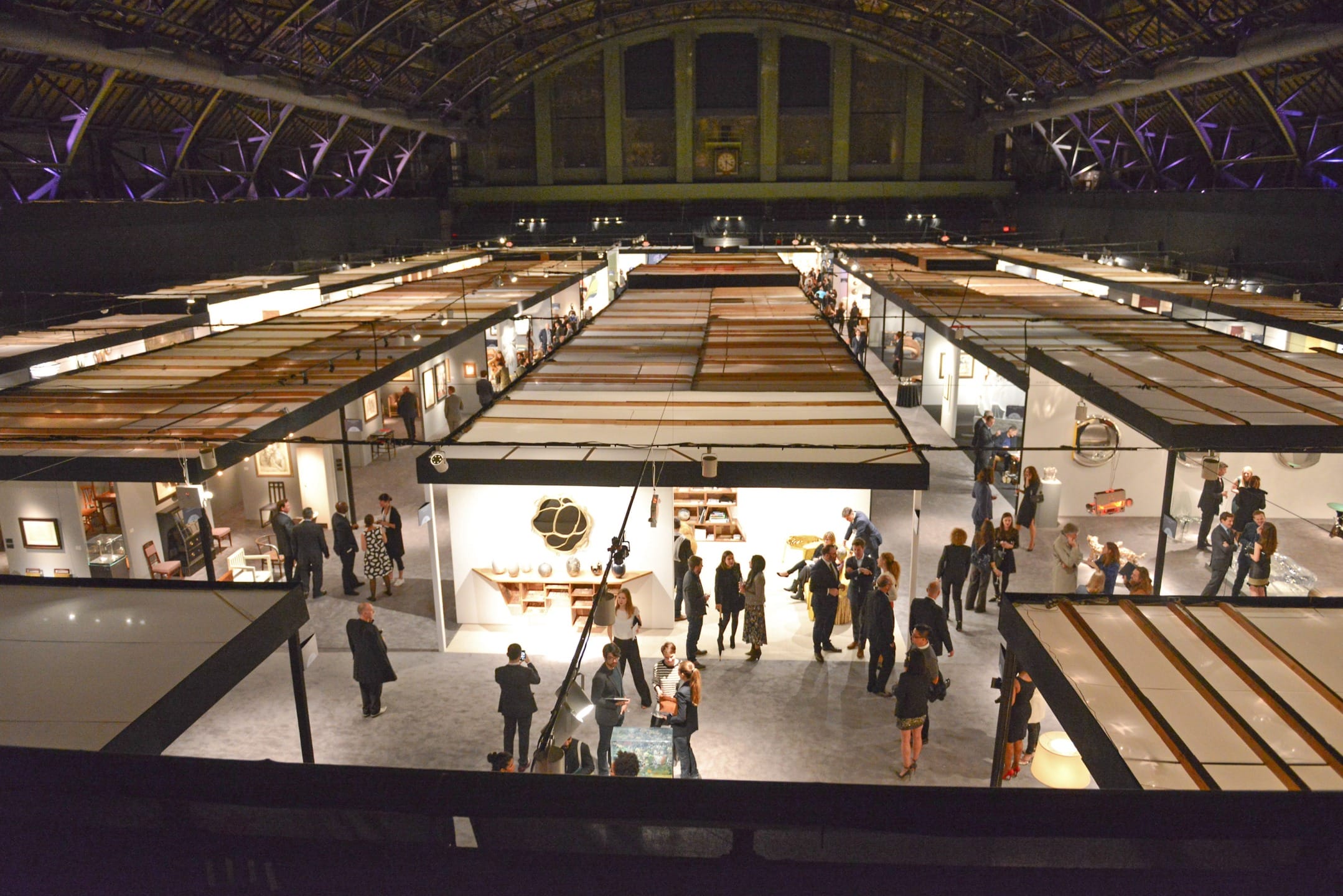 Art exhibition hall with people viewing various artistic displays and installations under a high ceiling.