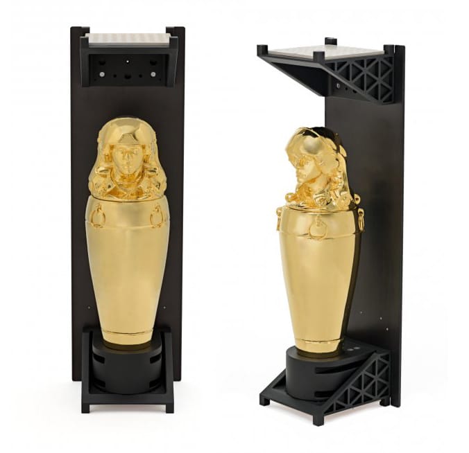 Golden urn in black display case with intricate, sculpted facial details and vertical design orientation.