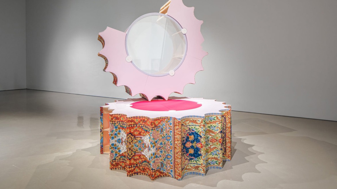 Contemporary art sculpture with a circular mirror, pink and ornate patterned elements, displayed in a modern gallery.