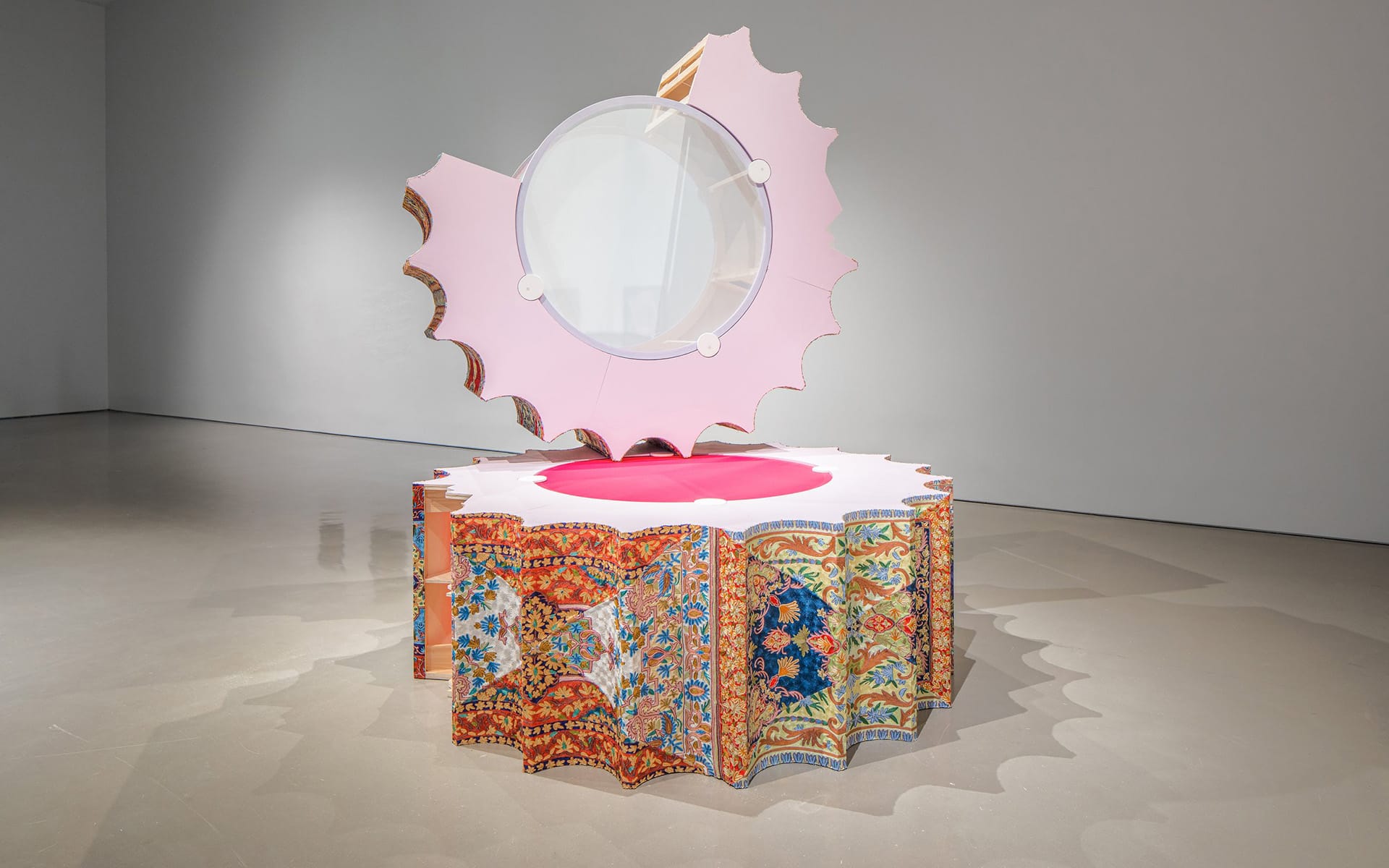 Contemporary art sculpture with a circular mirror, pink and ornate patterned elements, displayed in a modern gallery.