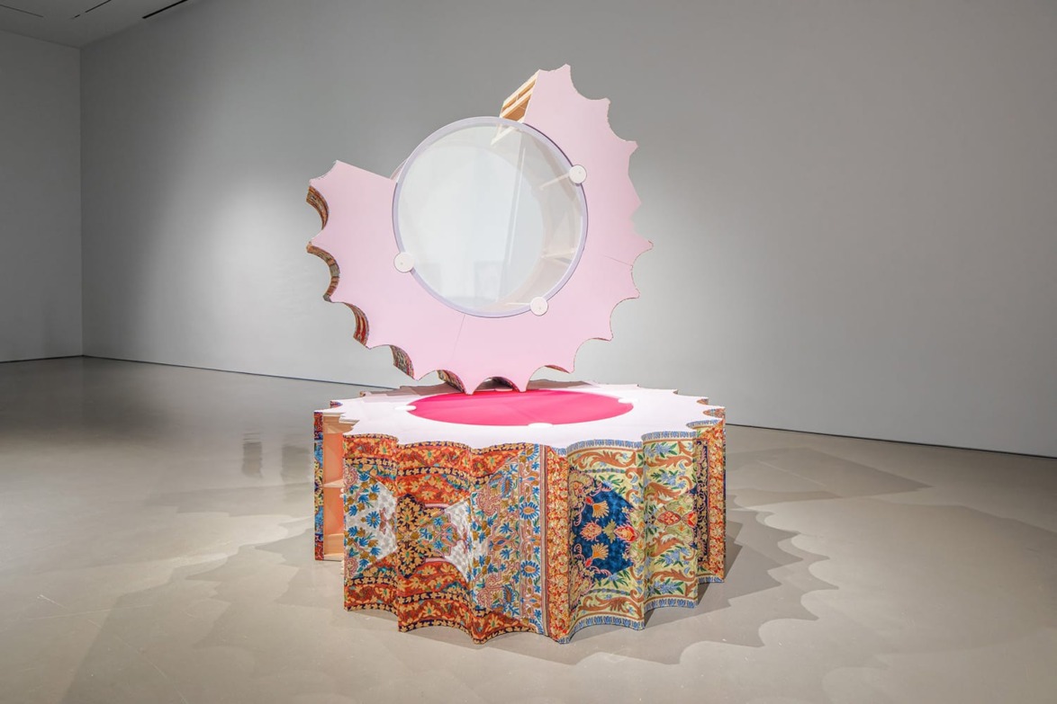 Colorful abstract sculpture with intricate patterns, featuring a circular mirror and scalloped pink edges in a gallery setting.