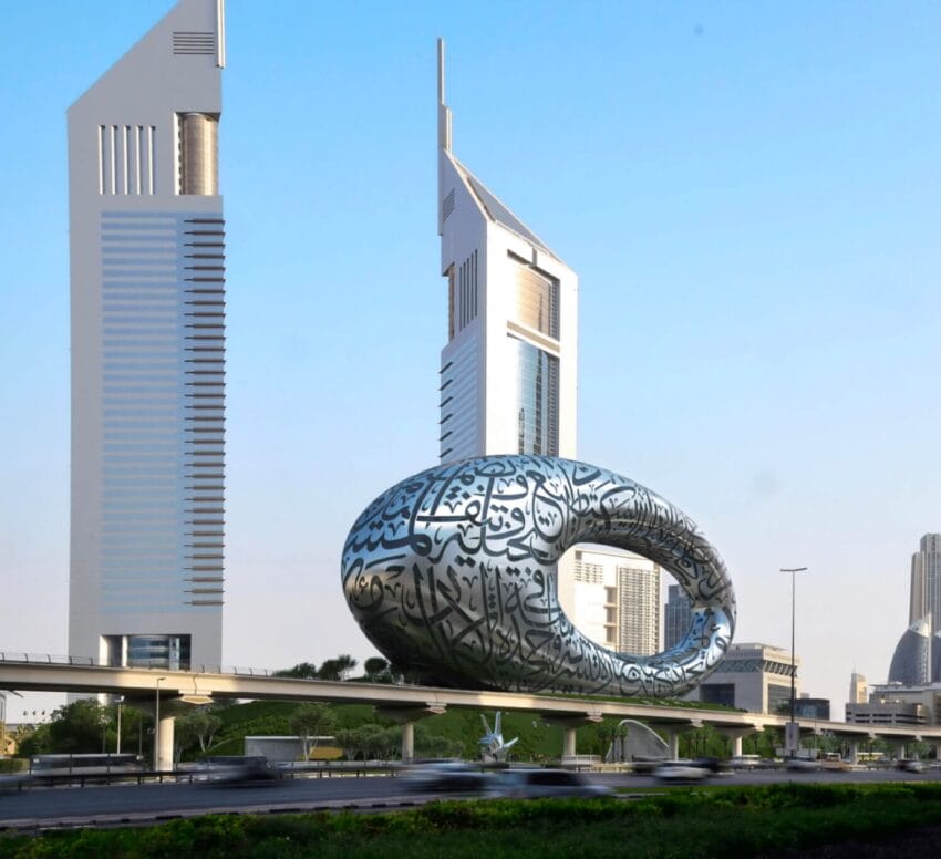 Futuristic silver building with Arabic calligraphy in Dubai, set against a clear blue sky and tall skyscrapers in the background.