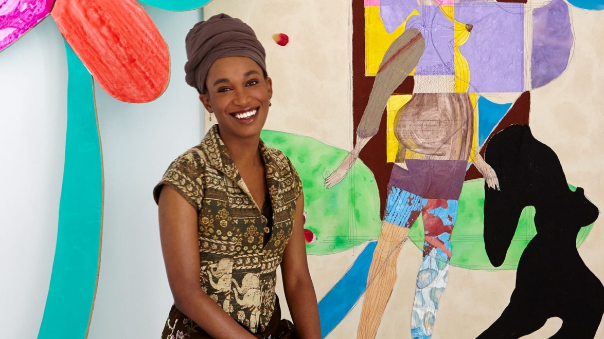 Smiling woman in patterned dress and headwrap standing beside colorful abstract painting.