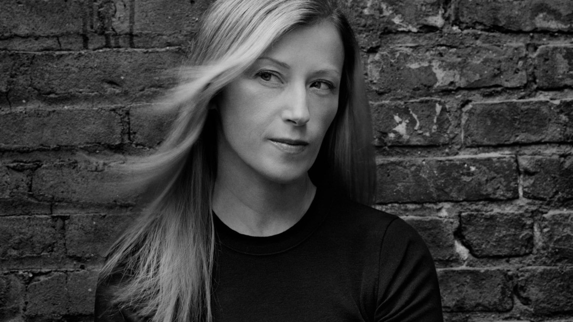 Woman with long hair standing against a brick wall, looking sideways in a black and white photo.