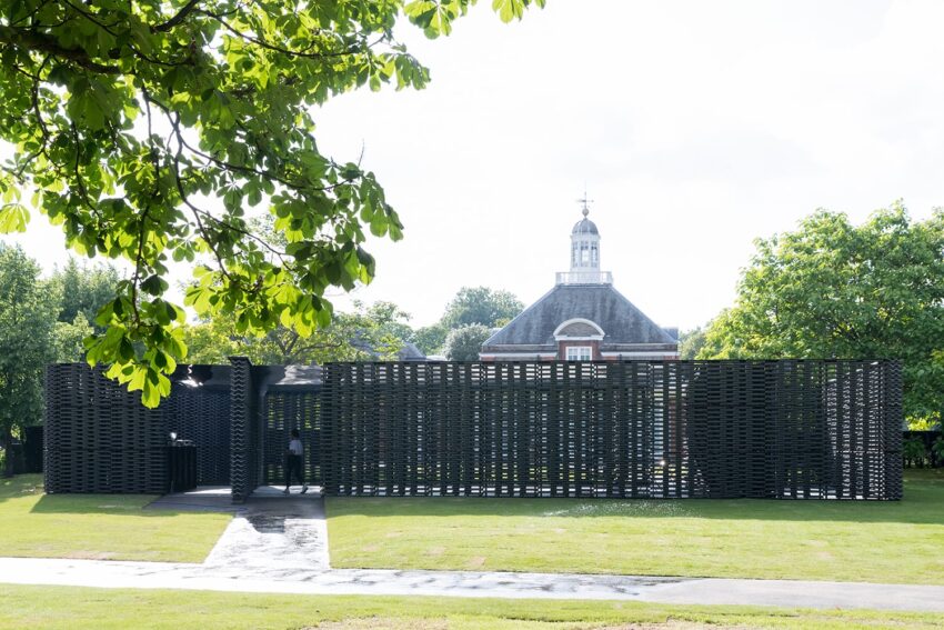 Black pavilion with woven texture in a green park setting, bright day, trees and historical building in the background.