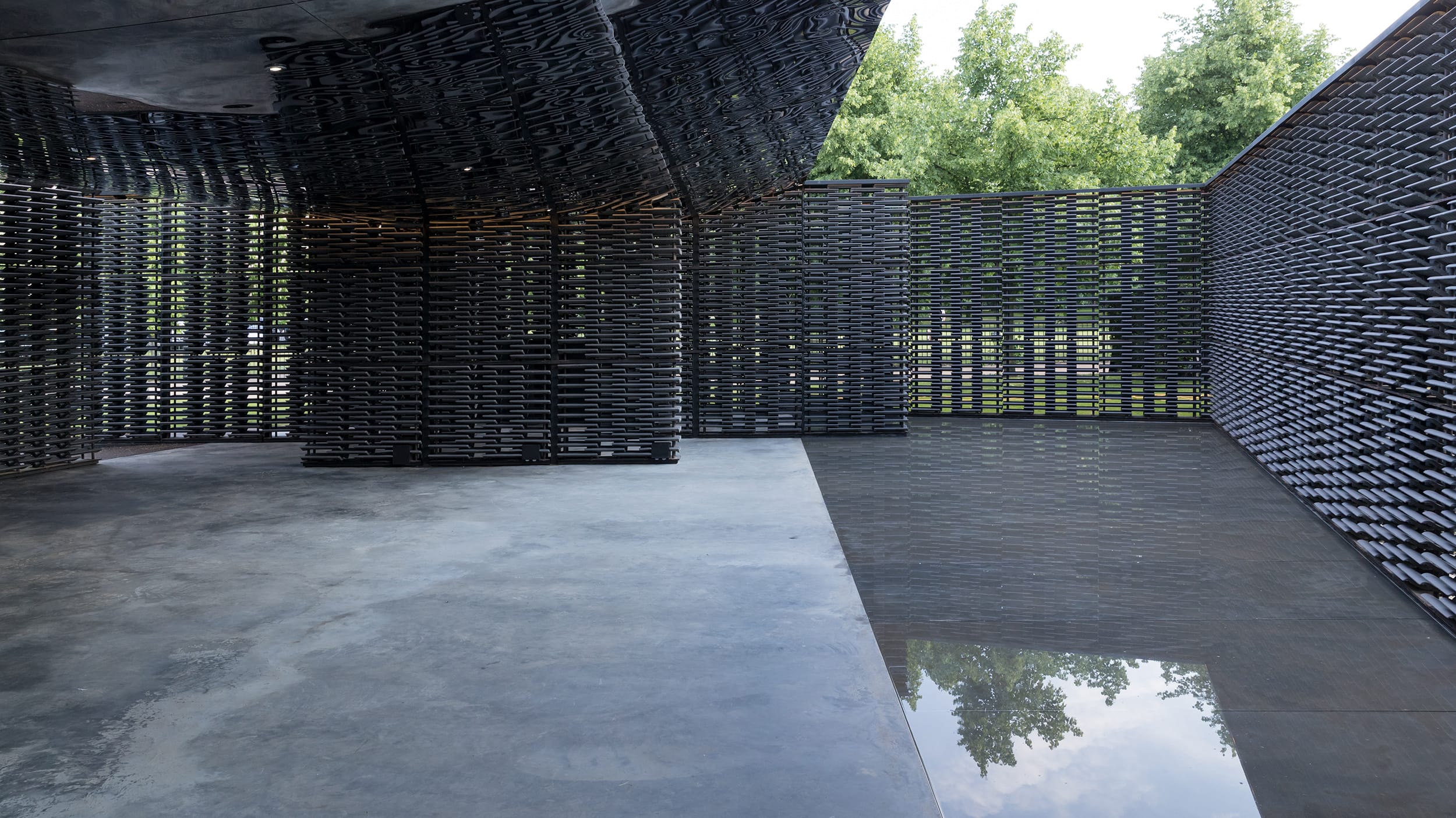 Modern architectural structure with black lattice walls and polished concrete floor, surrounded by greenery.