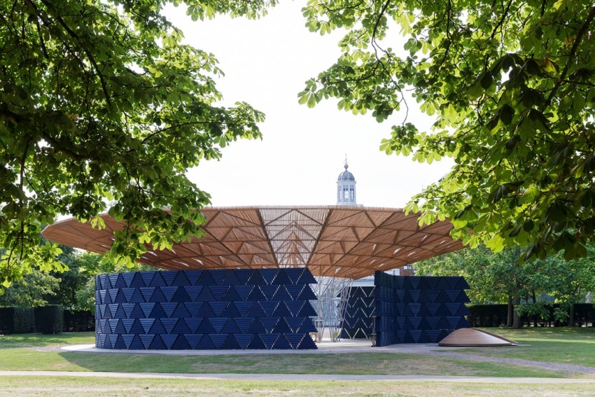 Modern pavilion with geometric design and wooden roof, surrounded by lush greenery and a historic building in the background.