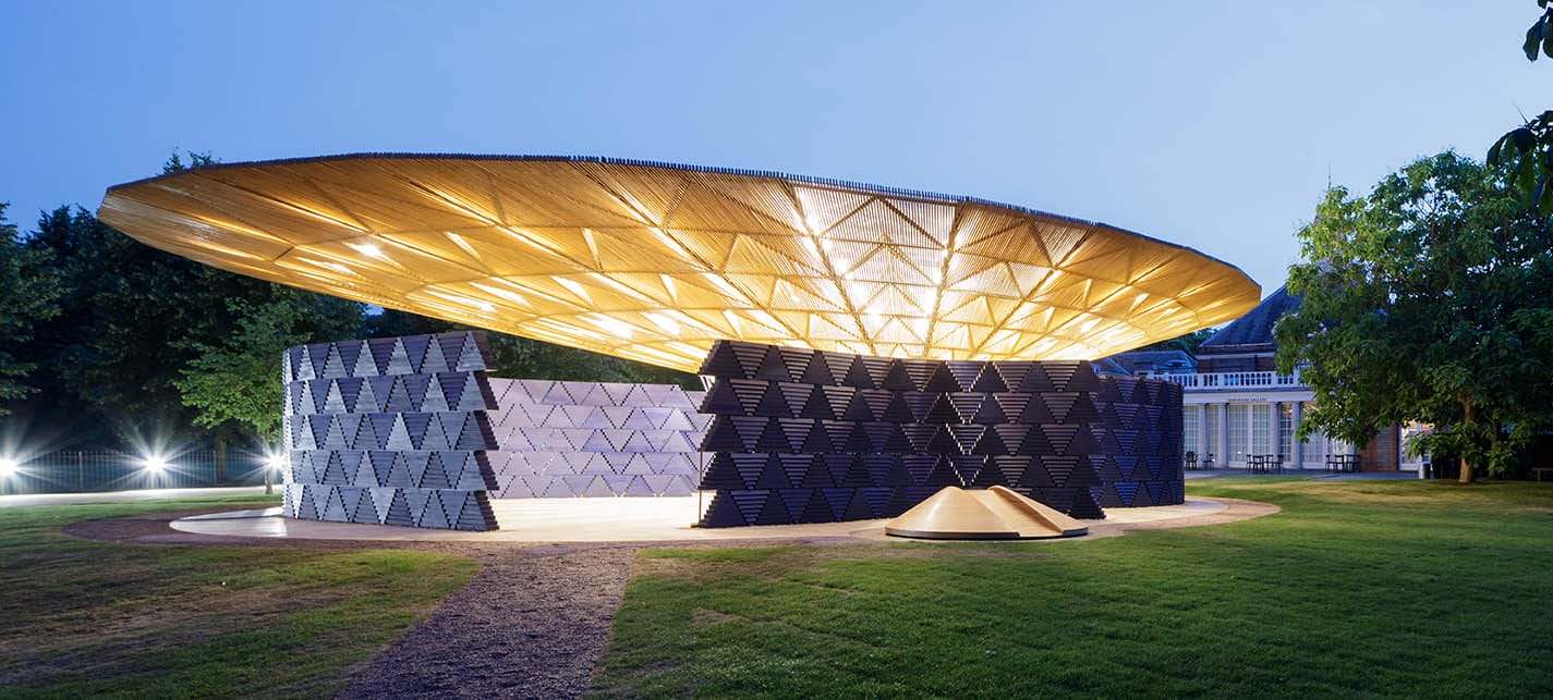 Circular architectural pavilion with illuminated roof, surrounded by green lawn and contemporary geometric walls at dusk.