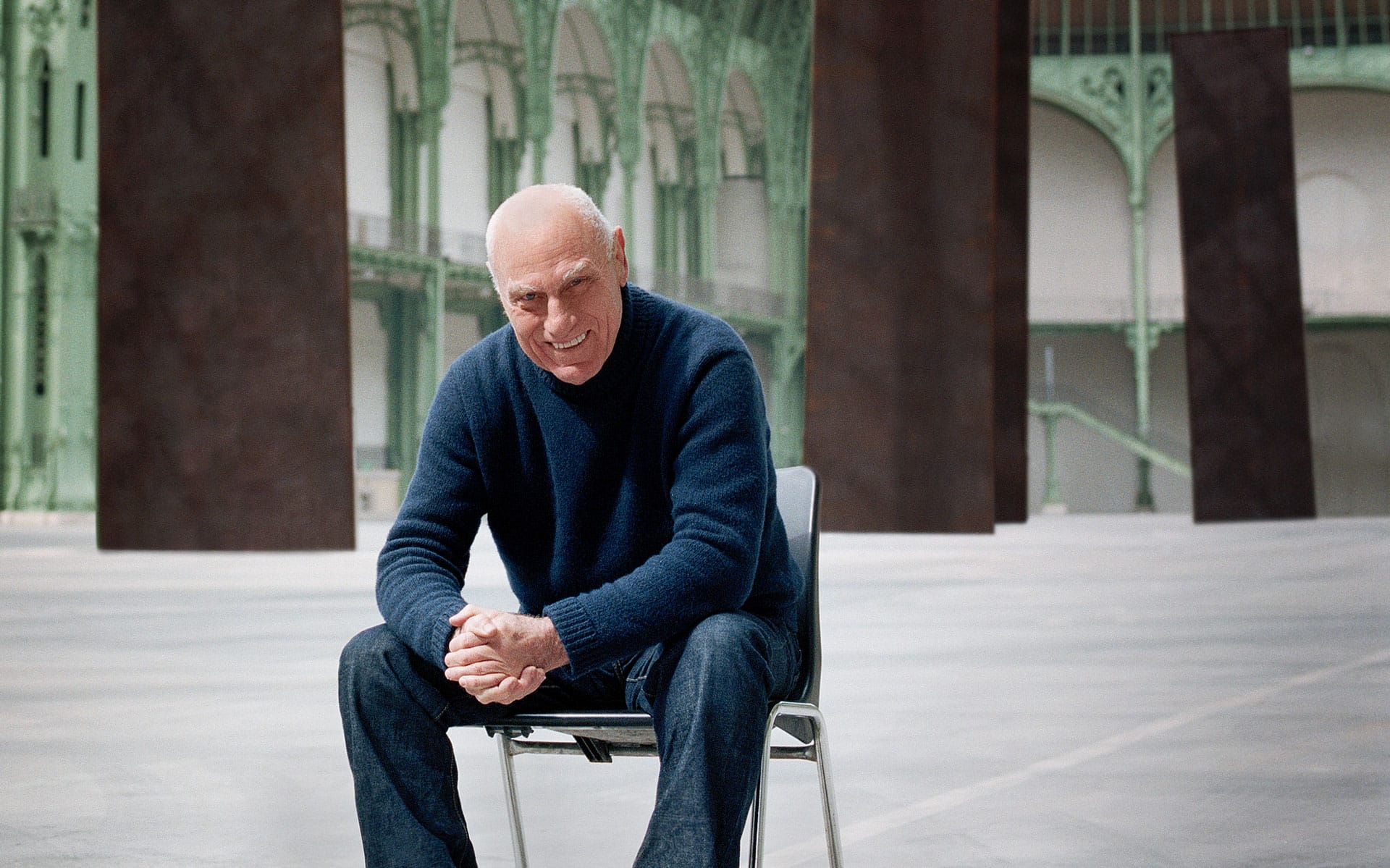Man sitting on a chair in a spacious room with tall, abstract art pieces and intricate architectural details in the background.