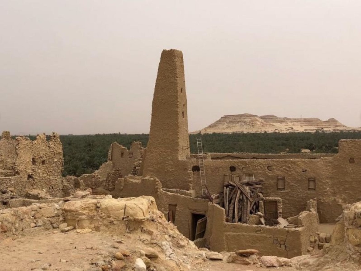 Ancient desert ruins with tall tower, surrounding stone walls, and distant mountain under a hazy sky.