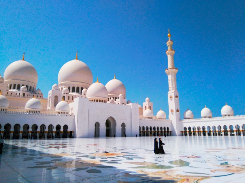Grand mosque with large white domes and minaret under clear blue sky, two people walking in the foreground.