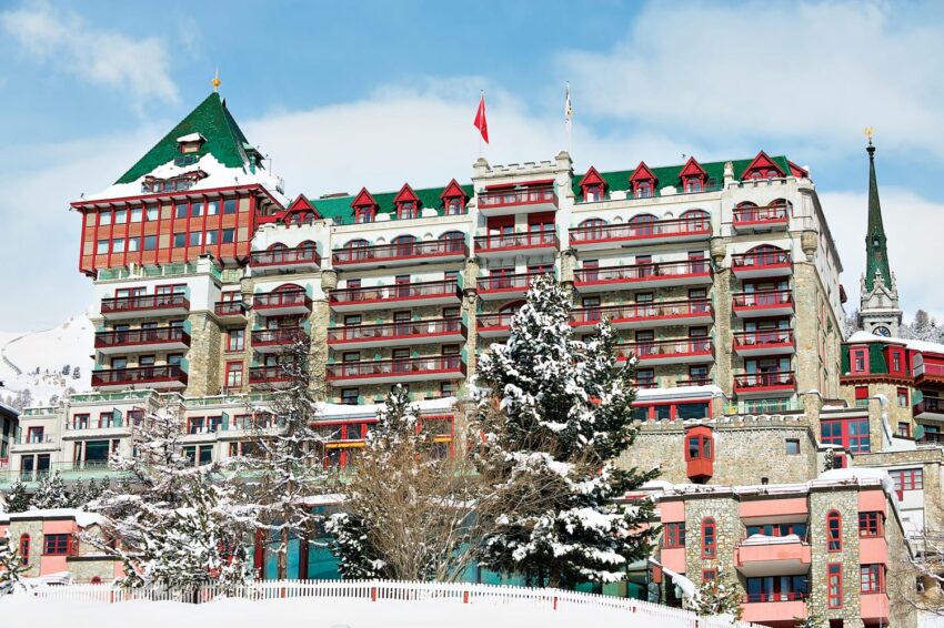 Historic hotel with red accents surrounded by snowy trees and mountains under a clear blue sky.