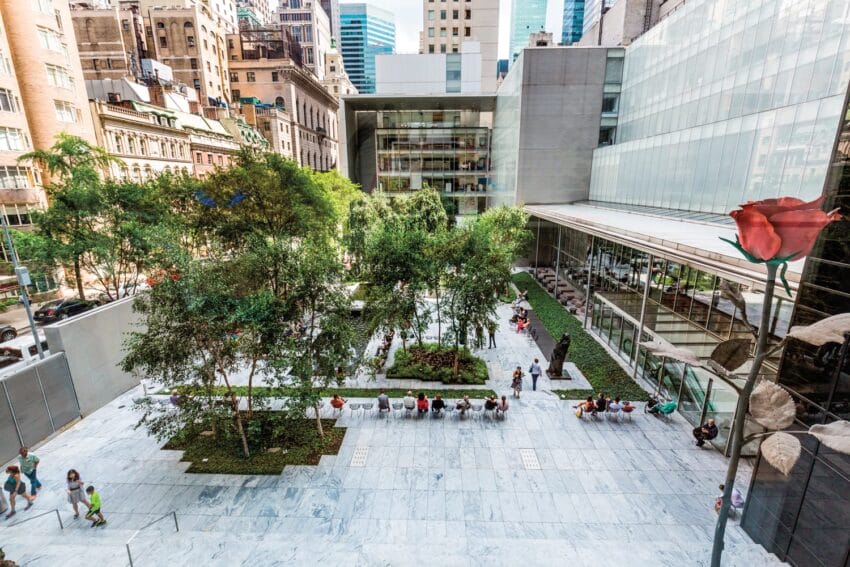Museum courtyard with trees, sculptures, and people sitting, surrounded by tall buildings in a busy urban setting.