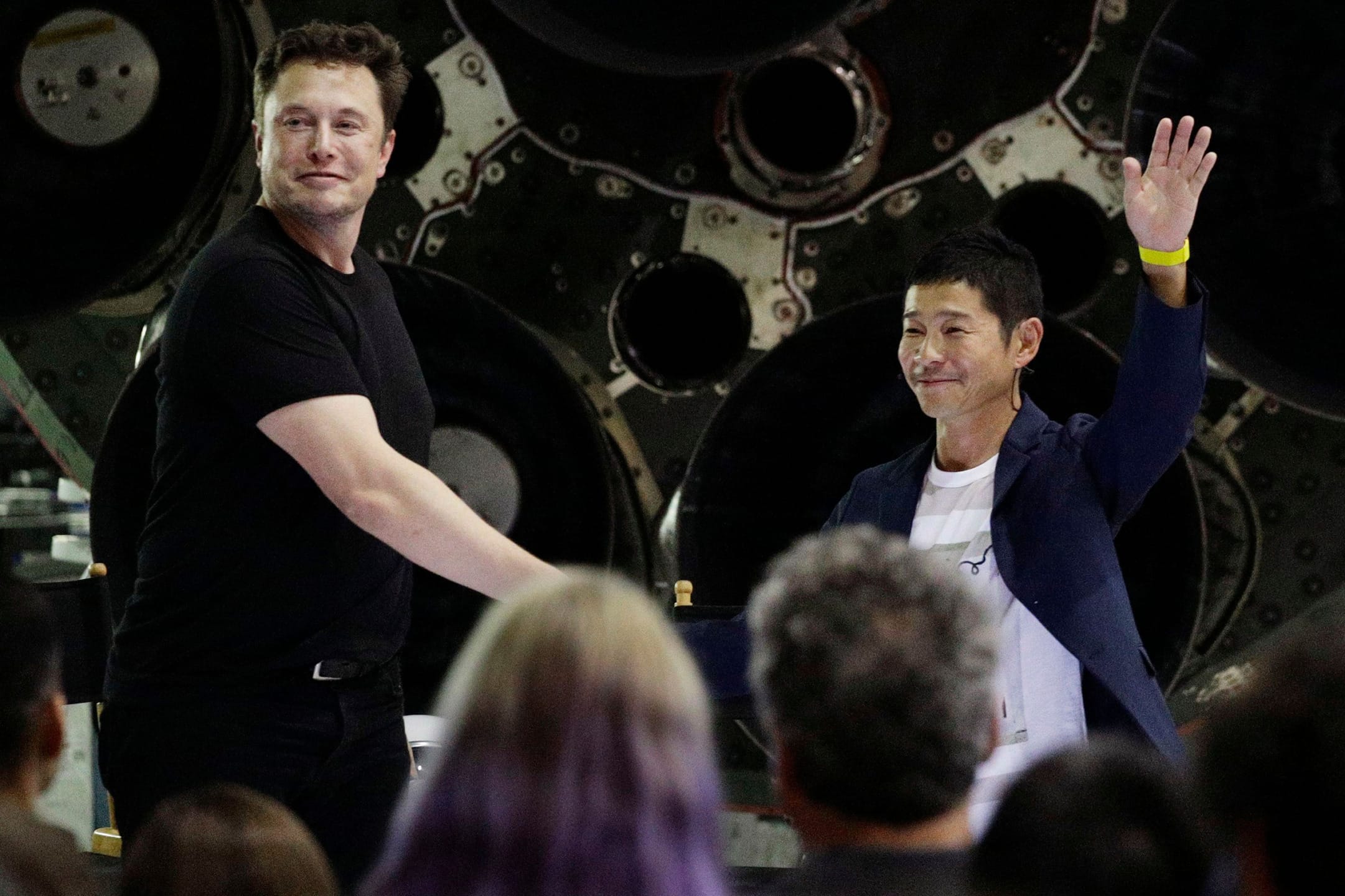Two men standing on stage, with one waving to an audience, in front of a large industrial backdrop.