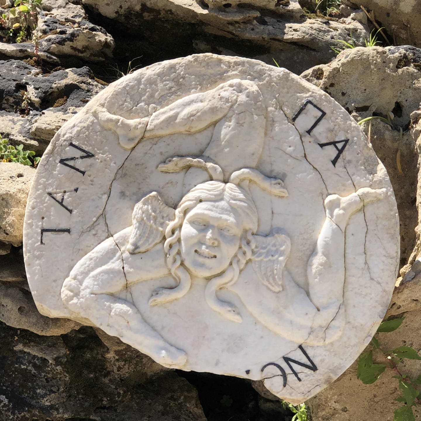 Ancient stone carving depicting a Gorgon face surrounded by three intertwined legs on a round plate with Greek letters.