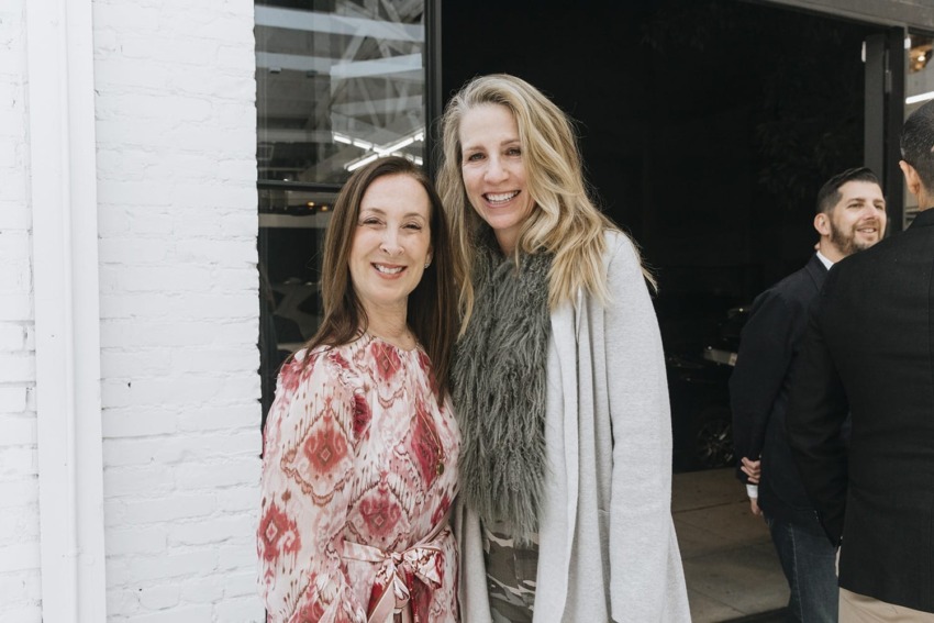 Two women smiling and posing together at an event with people and a building in the background.
