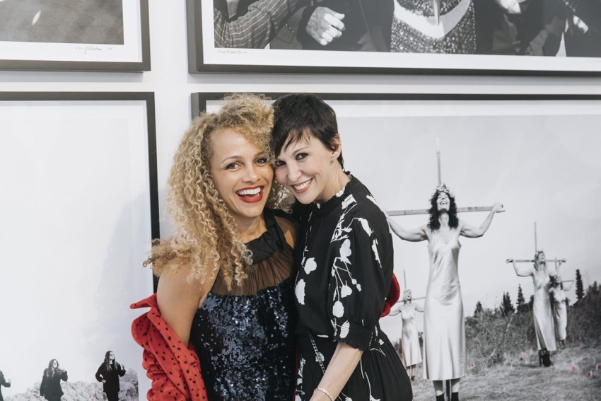 Two people smiling in front of black-and-white framed photographs on a wall.
