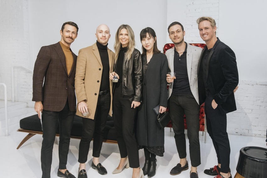 Group of six people dressed in fashionable attire standing together in a stylish, modern setting with a couch in the background.