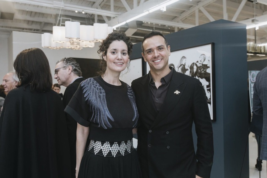 Two people posing and smiling at a gallery event with others socializing and artwork visible in the background.
