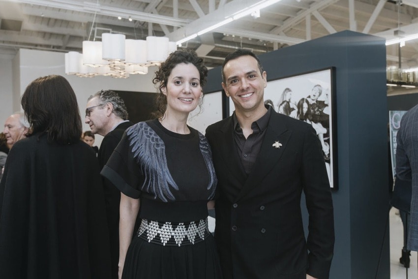 Two people posing and smiling at a gallery event with others socializing and artwork visible in the background.