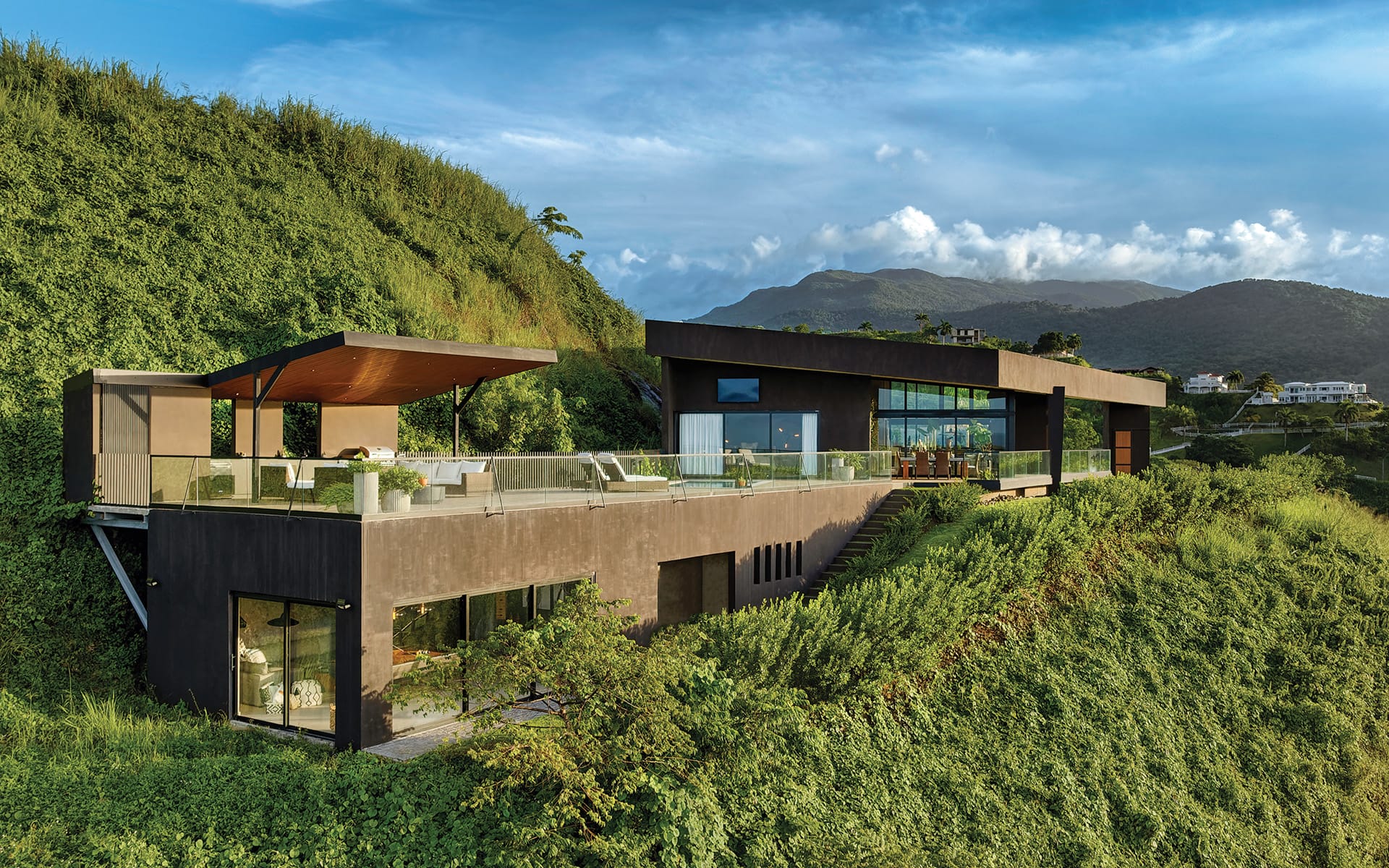 Modern hillside house with glass walls, surrounded by lush greenery and scenic mountain views under a blue sky.