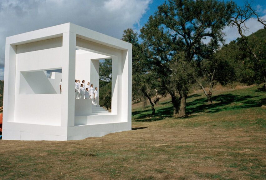 Group of people in white outfits standing inside a large, modern, open white cube structure in a grassy landscape with trees.