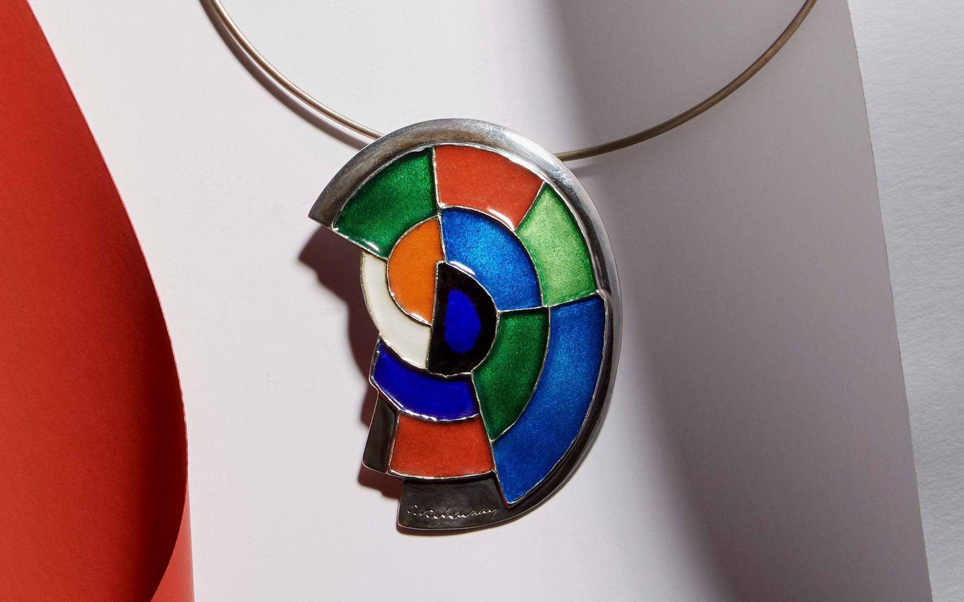 Colorful enamel and metal abstract pendant with geometric design on a necklace displayed against a red and white background.