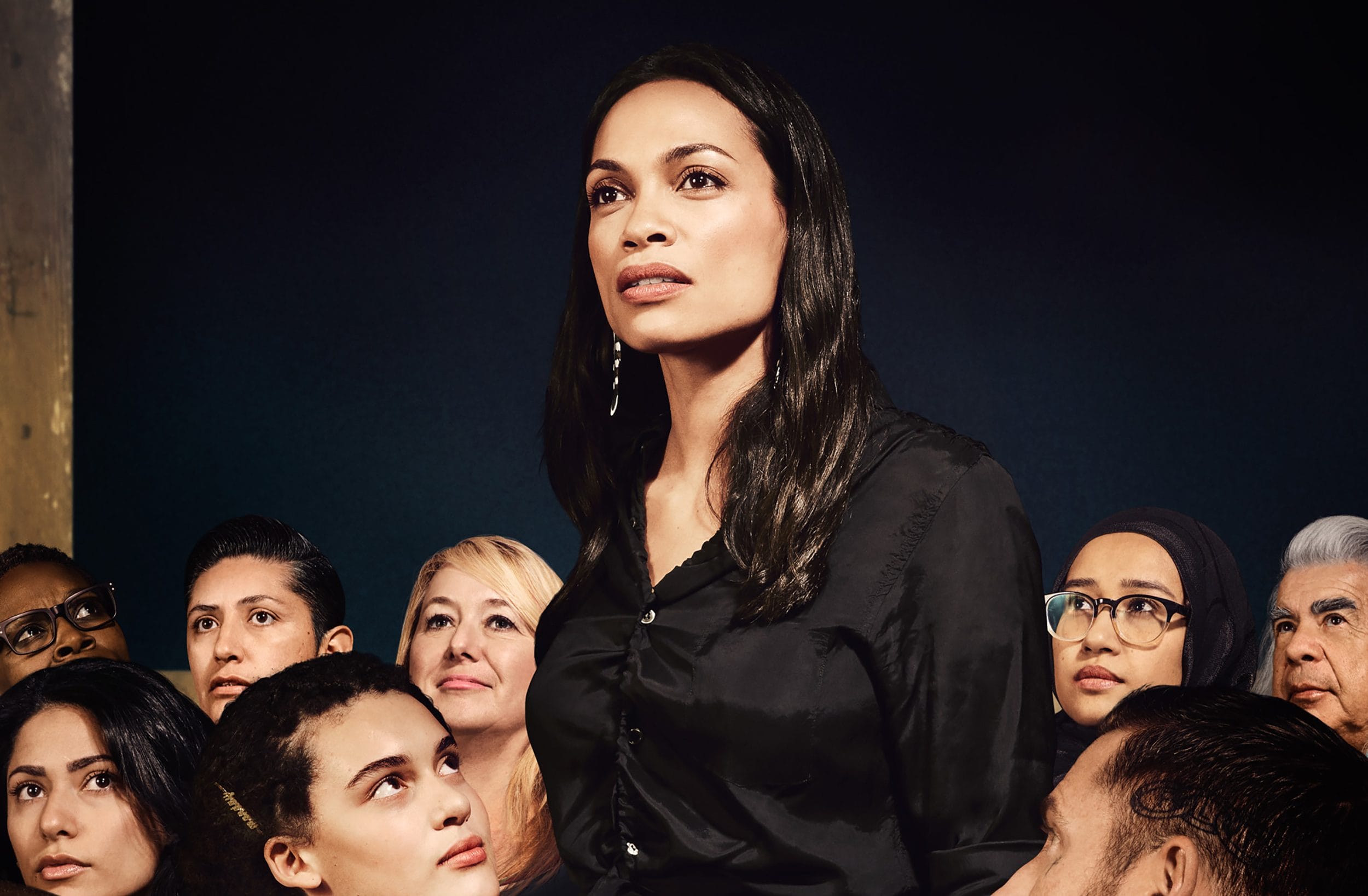 Woman in black shirt standing among a diverse group, all looking in different directions with a dark background.