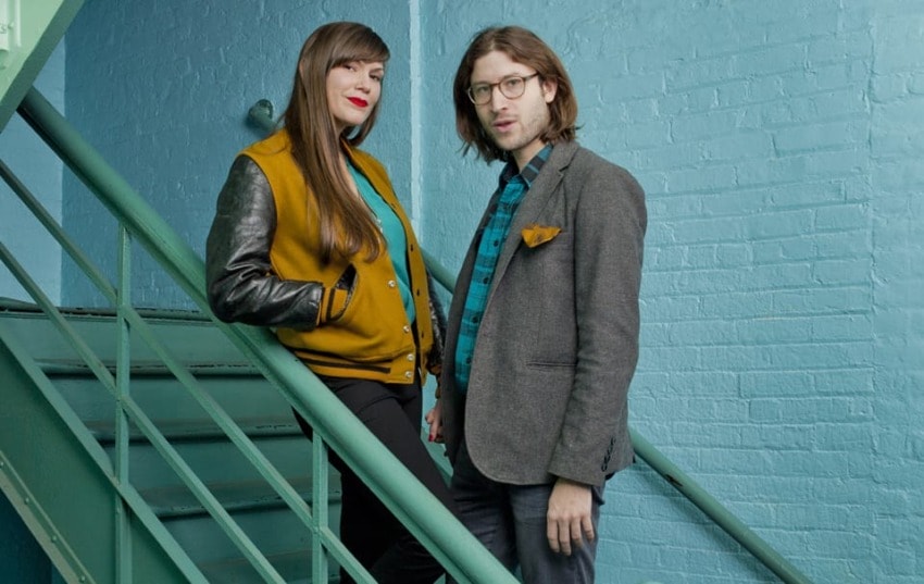 Two people standing on a staircase in a teal and mustard-themed setting.