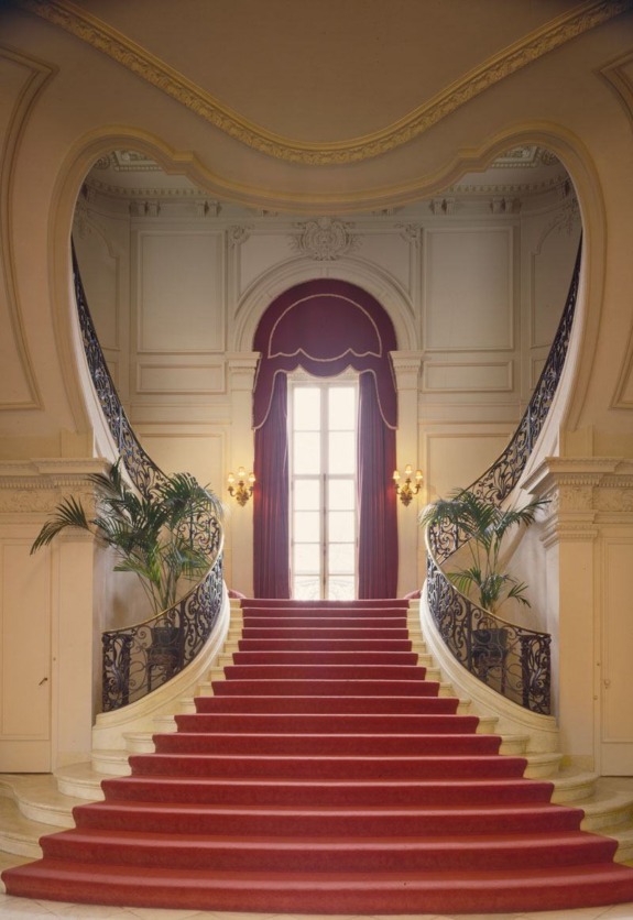 Elegant grand staircase with red carpet and ornate railing in a classic interior setting.