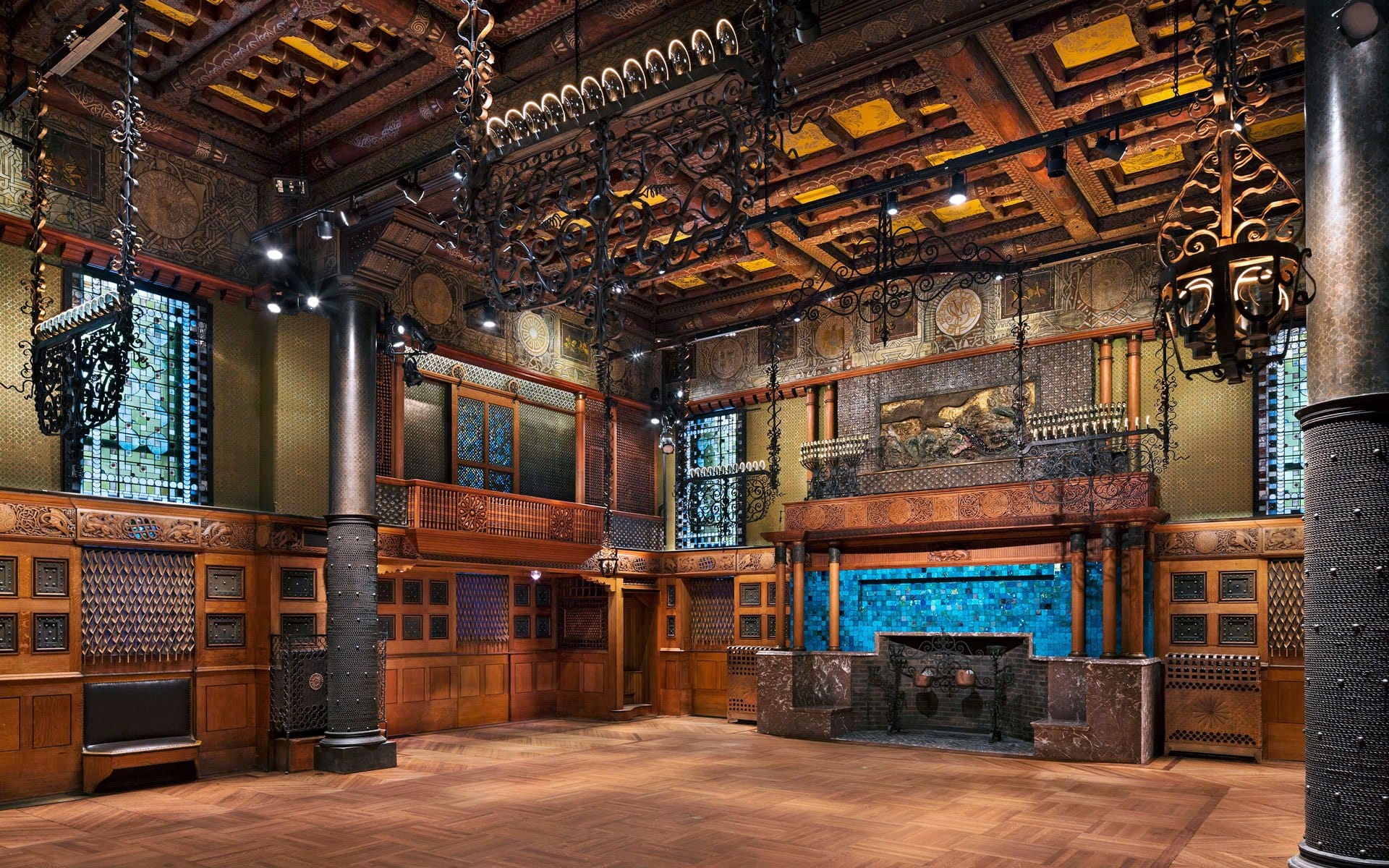 Ornate room with decorative wooden ceiling, intricate metalwork, stained glass windows, and a blue-tiled fireplace.