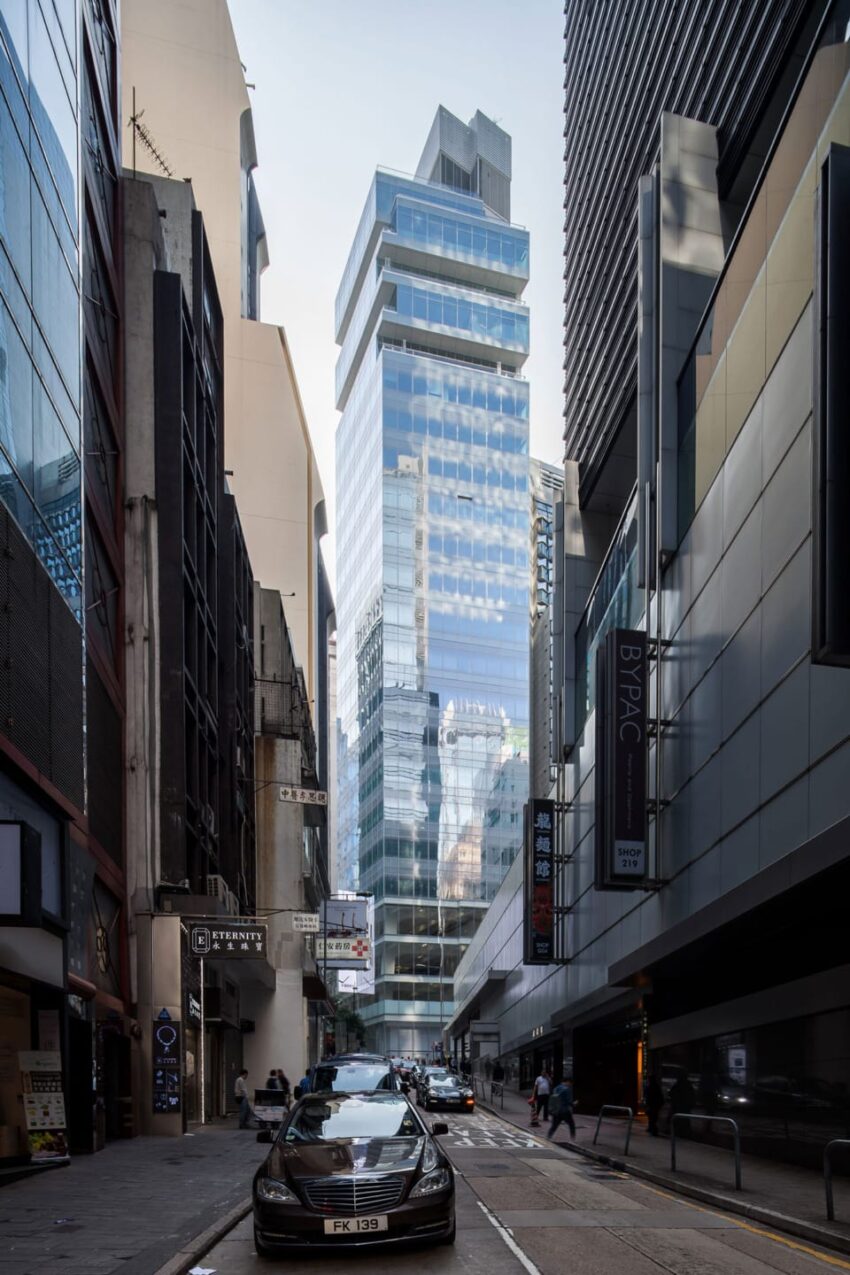 Modern glass skyscraper on a narrow city street with cars and pedestrians, surrounded by older buildings, under a blue sky.