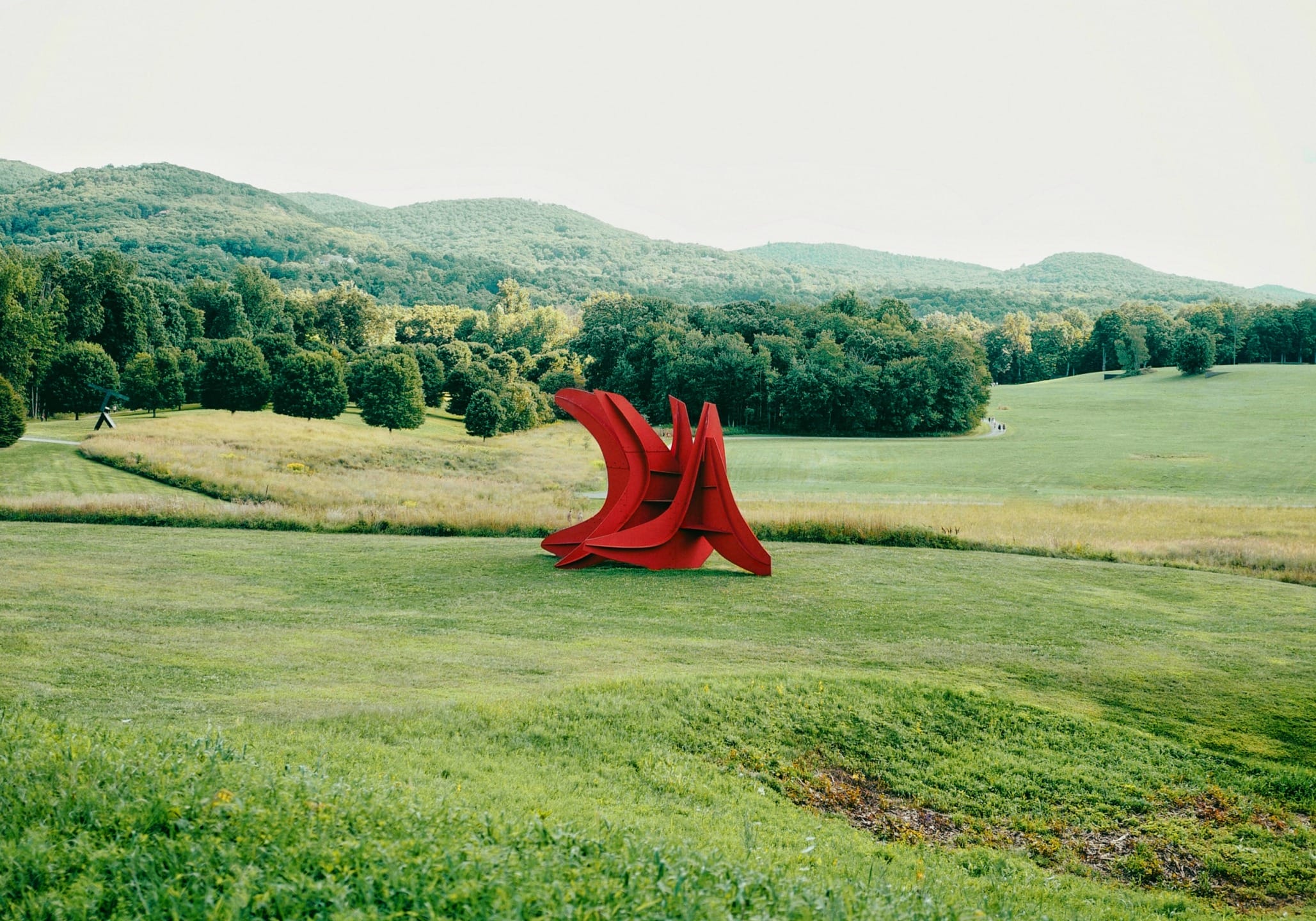 Red abstract sculpture on a grassy field with a backdrop of lush green hills and forest under a clear sky.
