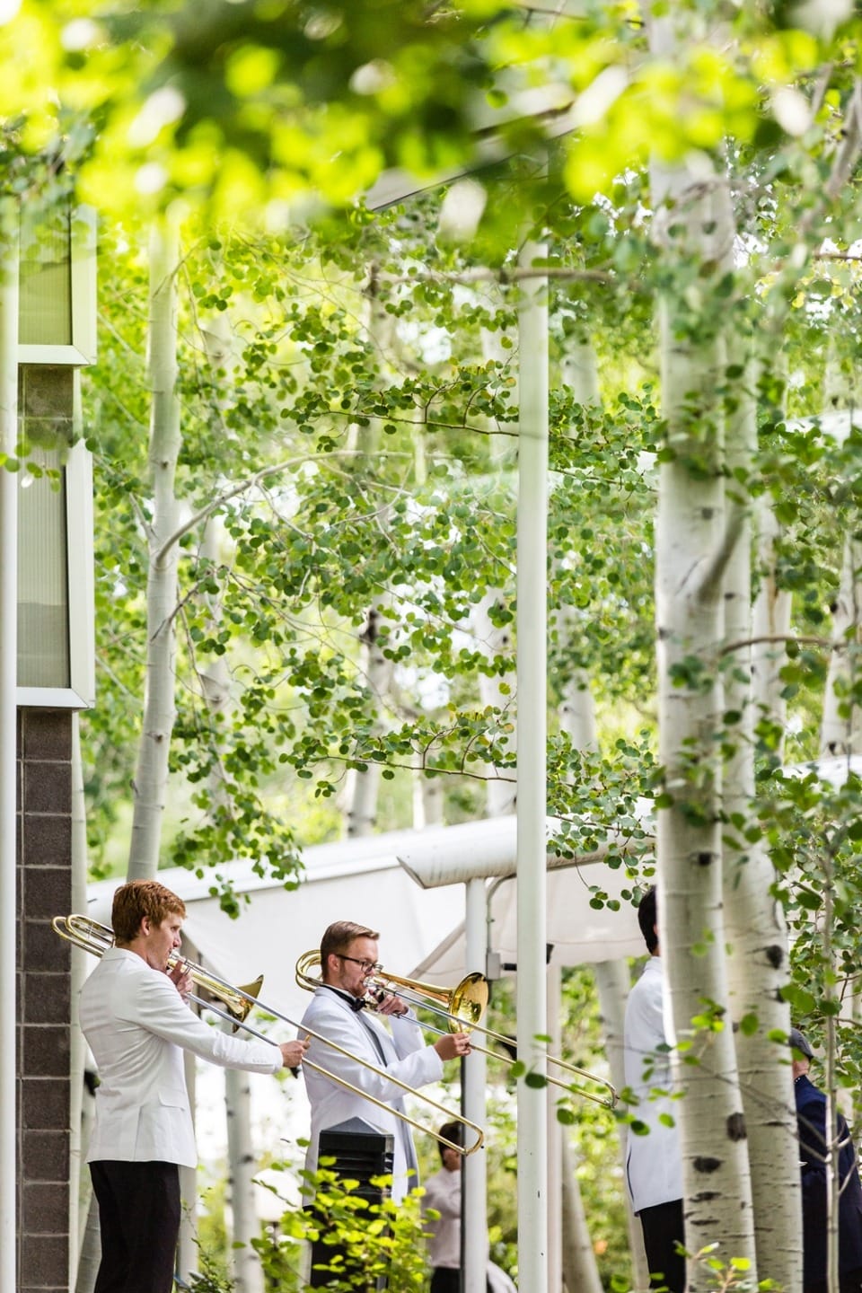 Musicians playing trombones outdoors among lush green trees in a sunny park setting