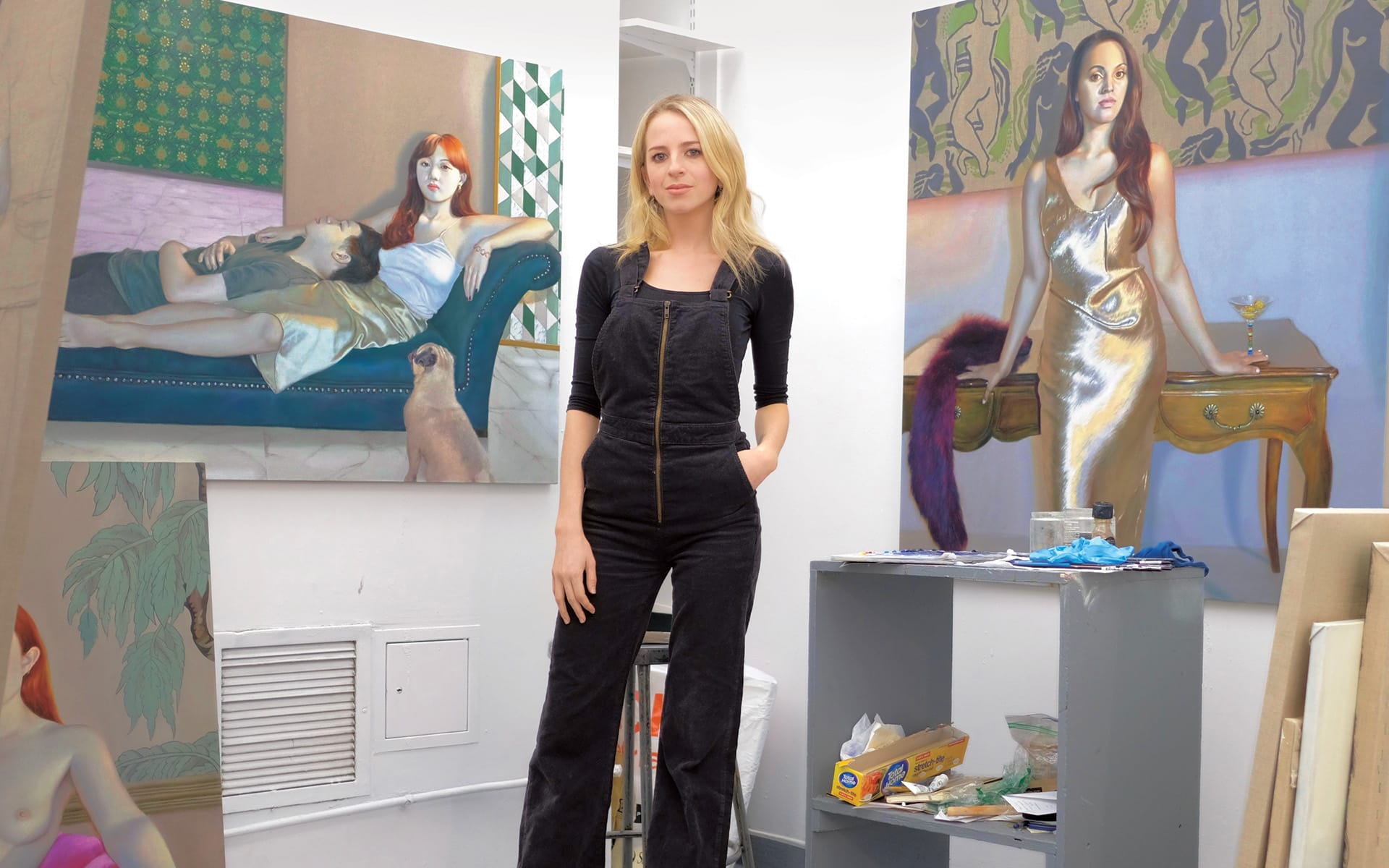 Artist standing in studio with paintings of women in elegant poses displayed on the walls and easel.