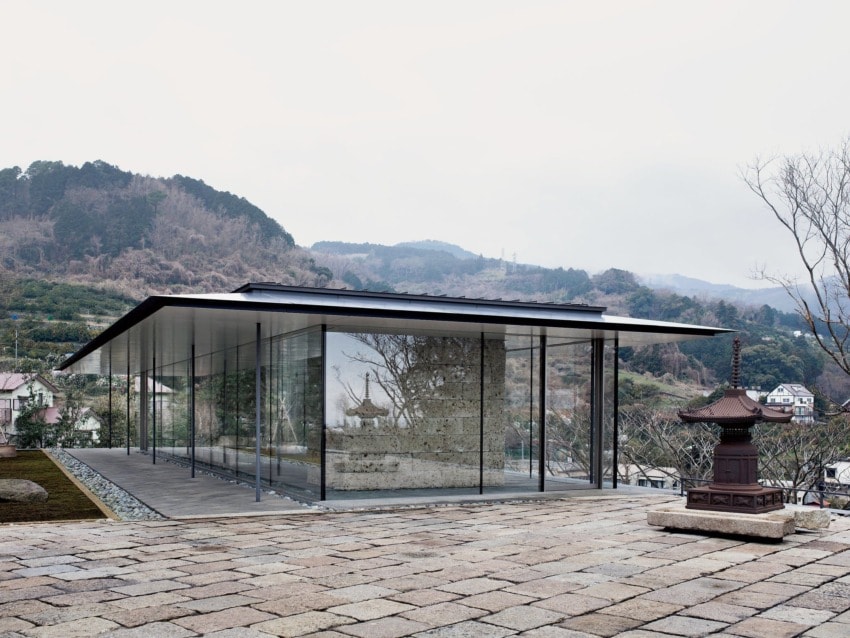 Modern glass building with reflection of mountains surrounded by trees and stone lantern, set in a mountainous landscape.