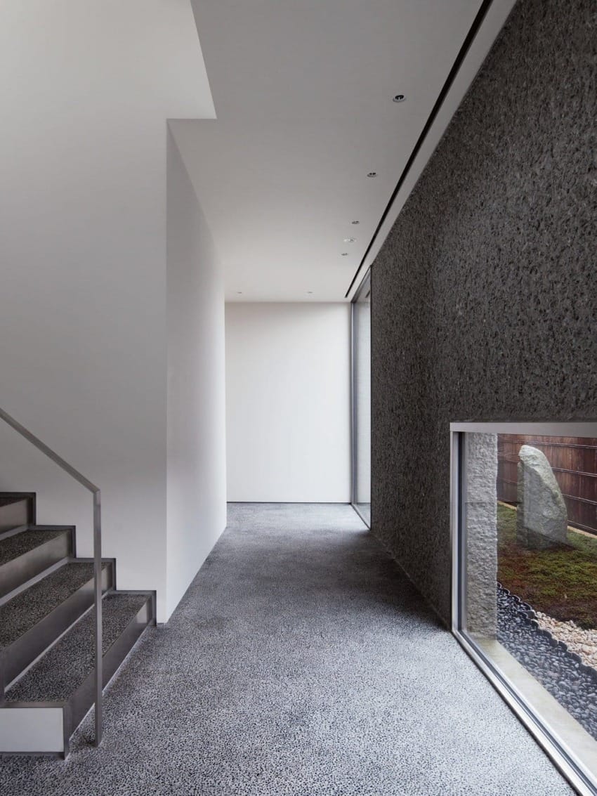 Modern minimalist hallway with pebble-textured flooring, white walls, and a window view of a small outdoor garden.