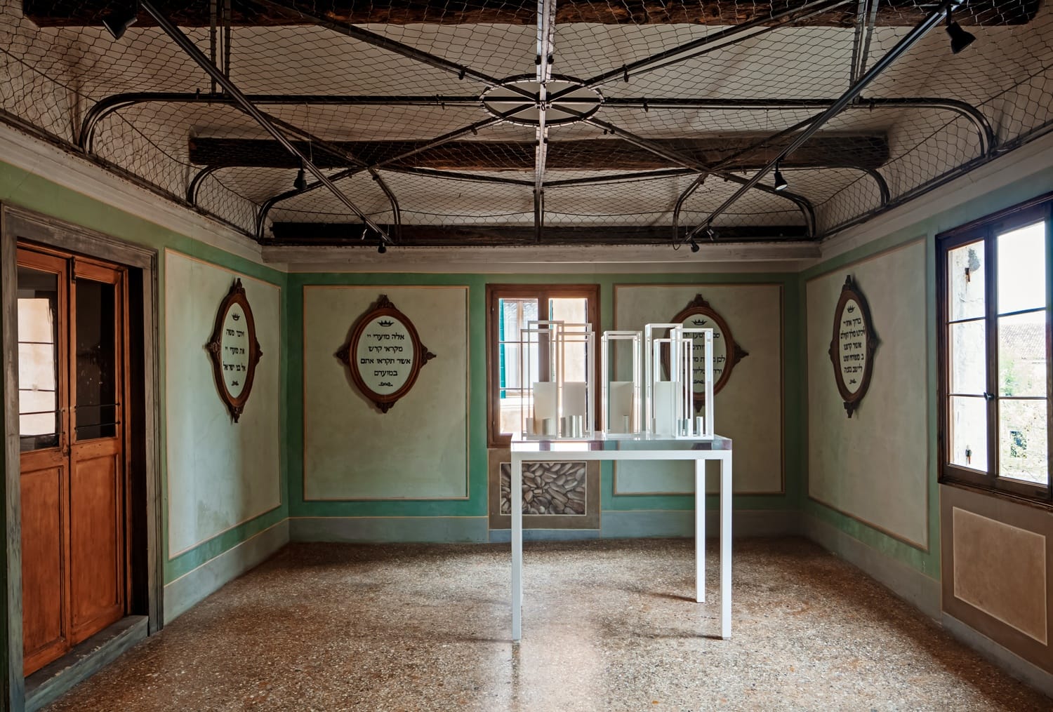Room with green walls, wooden beams, decorative cages on ceiling, and a modern art table display in the center.