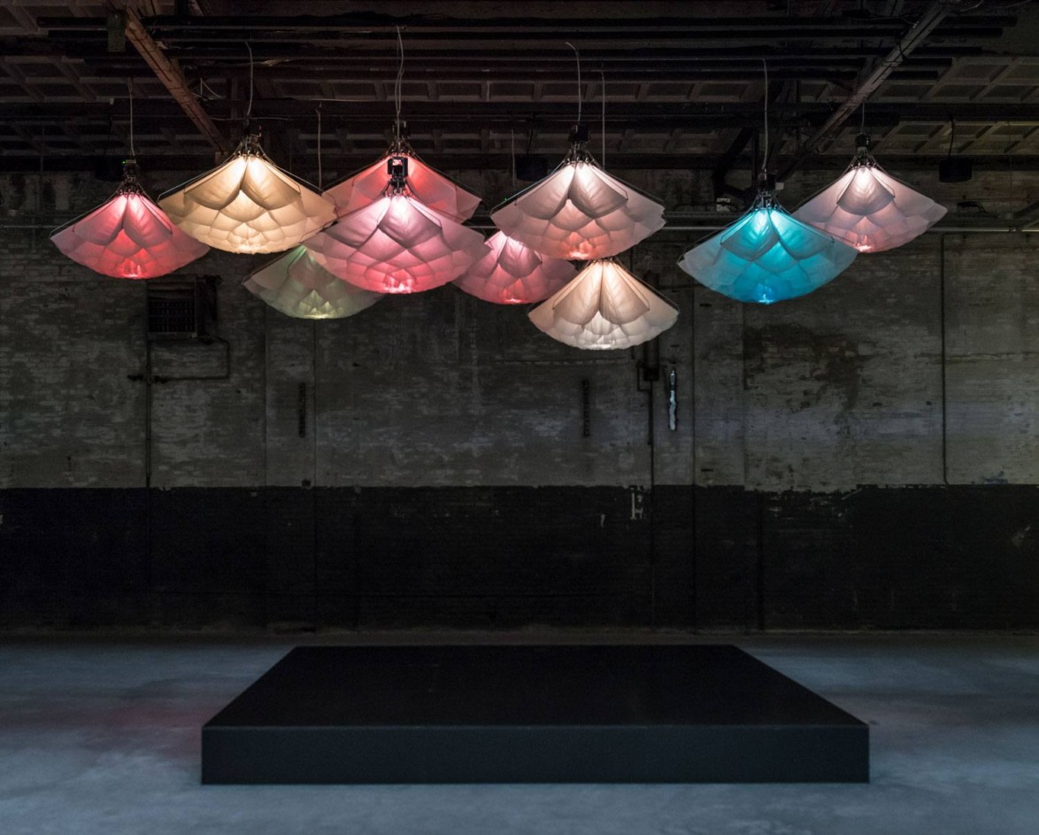 Colorful hanging light fixtures resembling fabric sails in a dimly lit industrial space with a platform below.