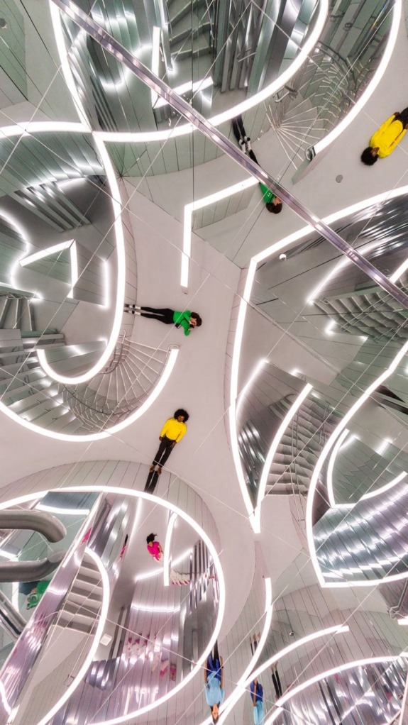 Futuristic mirrored room with multiple reflections of people in colorful clothing, creating an abstract pattern.