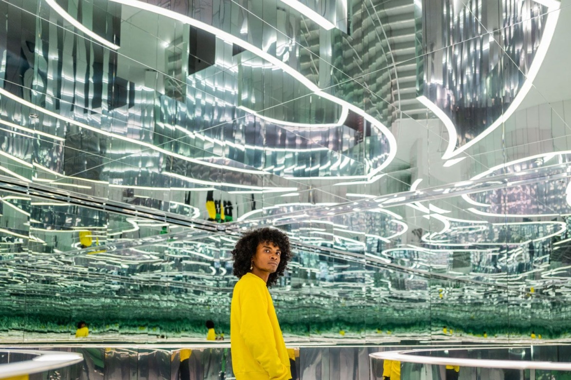 Person in yellow sweater stands in a room with mirrored walls and ceiling, reflecting vibrant patterns of light and color.