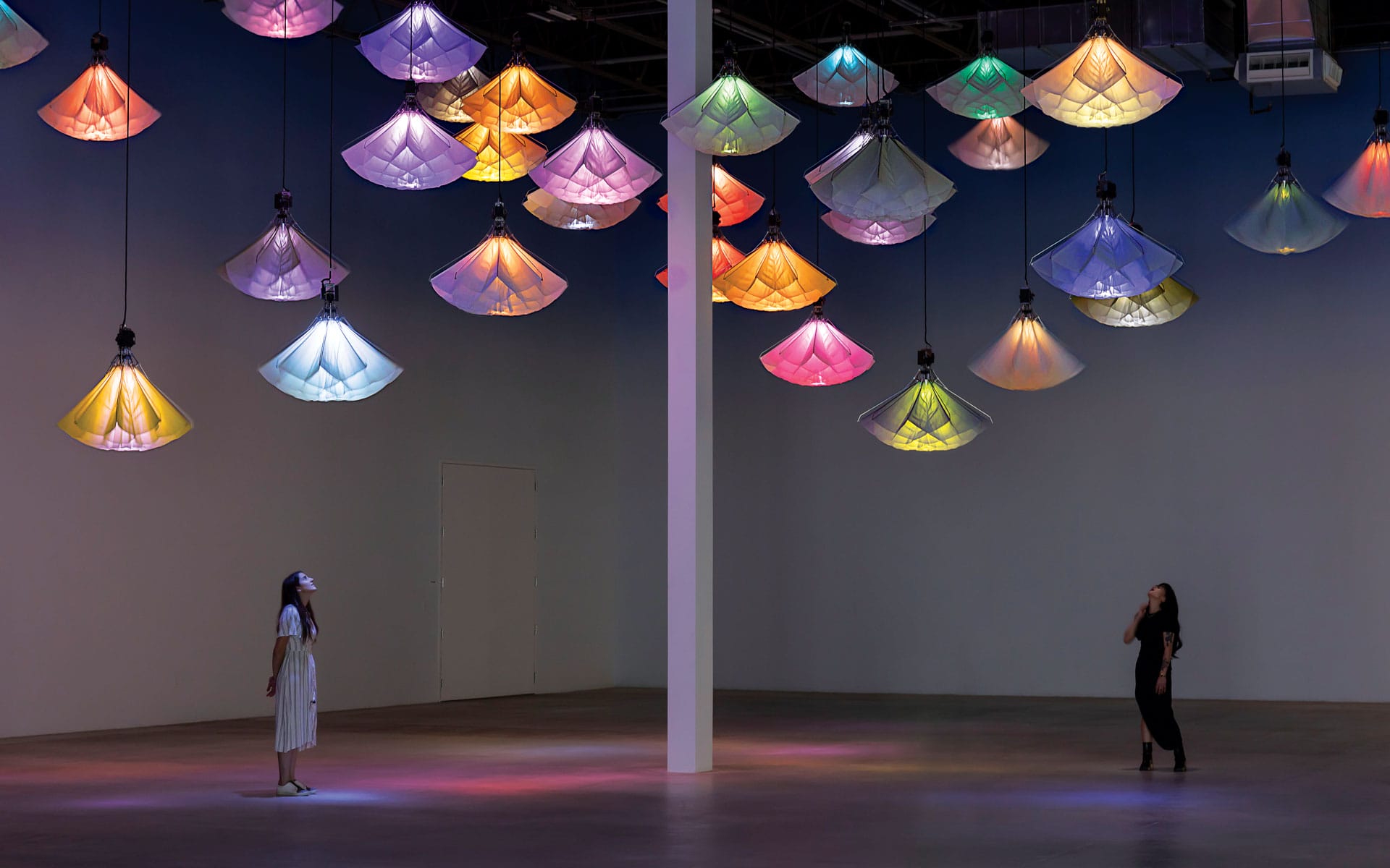 Two people admire colorful, hanging illuminated displays in a dark, open gallery space.